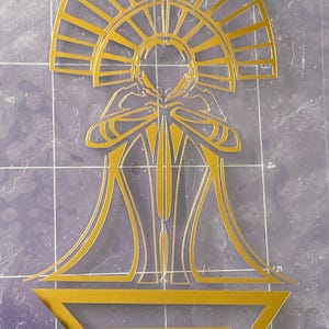 Puede incluir: Dise&ntilde;o dorado de estilo Art D&eacute;co con una figura central con halo, t&uacute;nicas fluidas y un tri&aacute;ngulo geom&eacute;trico en la base. El dise&ntilde;o est&aacute; cortado de una l&aacute;mina dorada.