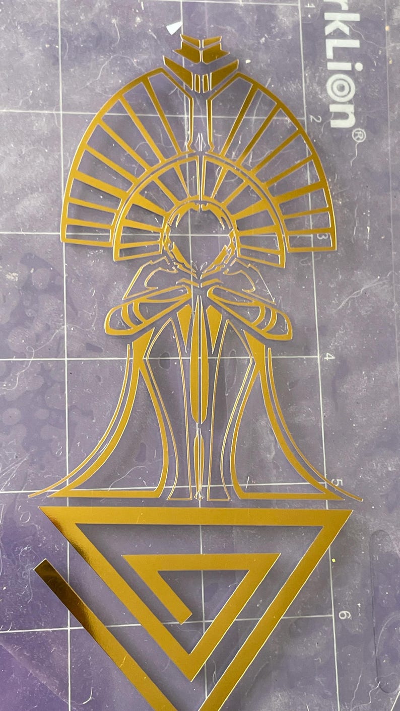 Puede incluir: Dise&ntilde;o dorado de estilo Art D&eacute;co con una figura central con halo, t&uacute;nicas fluidas y un tri&aacute;ngulo geom&eacute;trico en la base. El dise&ntilde;o est&aacute; cortado de una l&aacute;mina dorada.