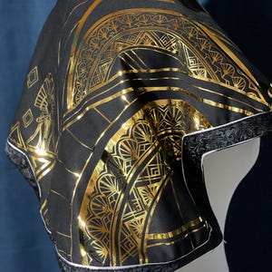 May include: Black cape with a gold Art Deco design. The cape features a repeating geometric pattern in gold foil. The edges are trimmed with black fabric and white piping. The cape is displayed on a white mannequin.