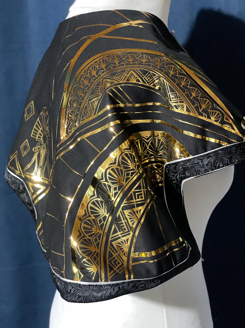 May include: Black cape with a gold Art Deco design. The cape features a repeating geometric pattern in gold foil. The edges are trimmed with black fabric and white piping. The cape is displayed on a white mannequin.