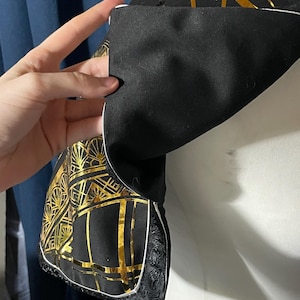May include: Close-up of a black fabric with a gold geometric pattern, possibly part of a garment. The design features gold lines and shapes on a black background. The fabric is held, revealing a white trim and decorative edge.