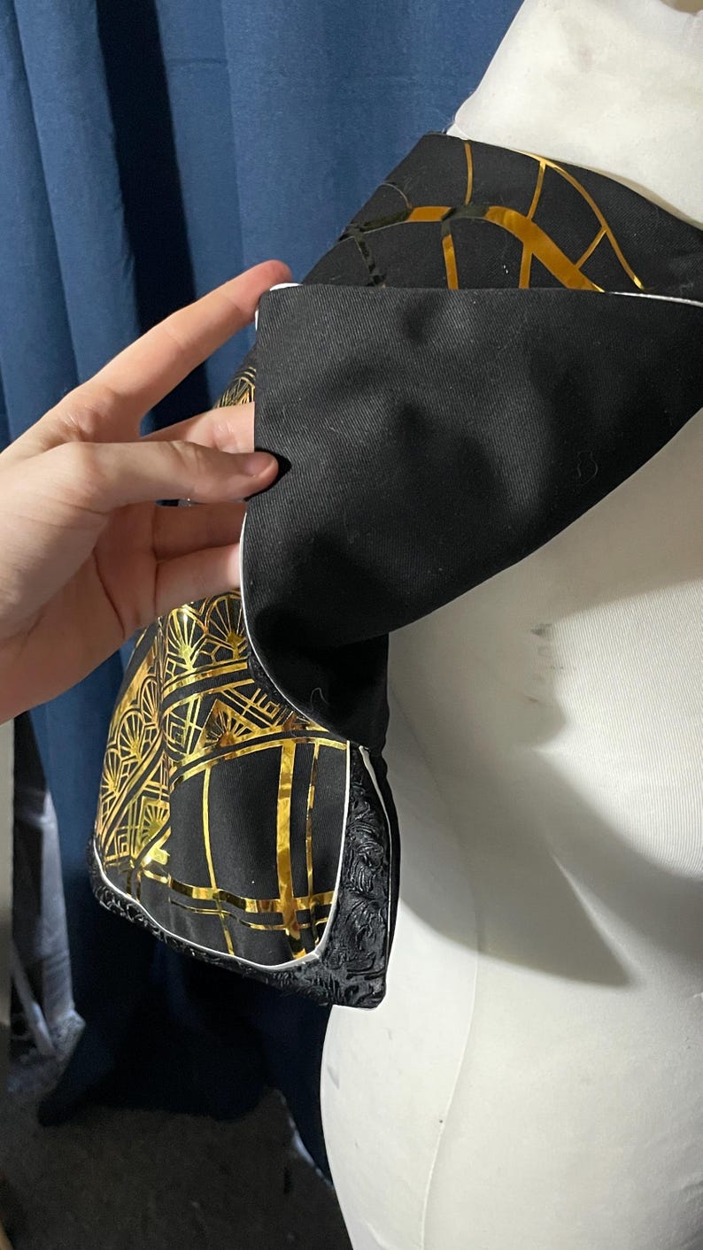 May include: Close-up of a black fabric with a gold geometric pattern, possibly part of a garment. The design features gold lines and shapes on a black background. The fabric is held, revealing a white trim and decorative edge.