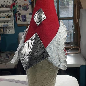 May include: A red conical hat with silver-colored decorative elements. The hat features a silver crest, diamond-shaped accents, and wing-shaped embellishments. The hat is displayed on a mannequin head, showcasing its design and craftsmanship.