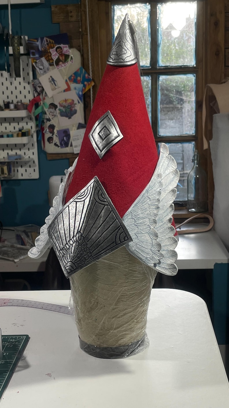 May include: A red conical hat with silver-colored decorative elements. The hat features a silver crest, diamond-shaped accents, and wing-shaped embellishments. The hat is displayed on a mannequin head, showcasing its design and craftsmanship.