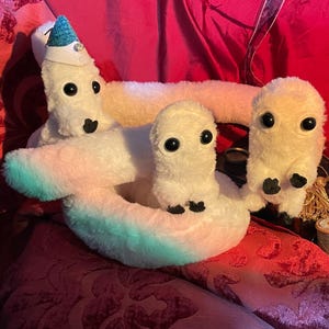 May include: Three white plush toys with large eyes. The toy in the center is wearing a blue and white party hat. The toys are arranged in a circle on a red fabric surface.