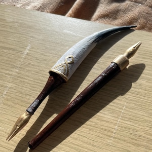 Puede incluir: Dos plumas estilogr&aacute;ficas de estilo antiguo. Una pluma presenta una pluma decorada en blanco y dorado con un mango marr&oacute;n oscuro. La otra pluma tiene un mango marr&oacute;n oscuro y una punta dorada. Ambas plumas est&aacute;n sobre una superficie de madera clara.