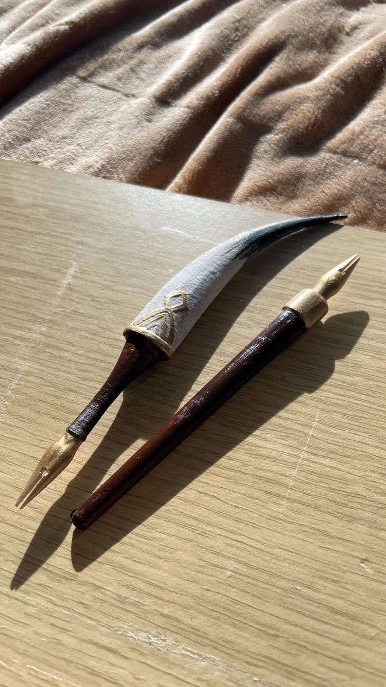 Puede incluir: Dos plumas estilogr&aacute;ficas de estilo antiguo. Una pluma presenta una pluma decorada en blanco y dorado con un mango marr&oacute;n oscuro. La otra pluma tiene un mango marr&oacute;n oscuro y una punta dorada. Ambas plumas est&aacute;n sobre una superficie de madera clara.