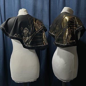 May include: Two black capes with gold geometric patterns and fringing are displayed on white mannequins. The capes have a short, rounded shape and are decorated with gold lines and shapes. The background is a dark blue curtain.