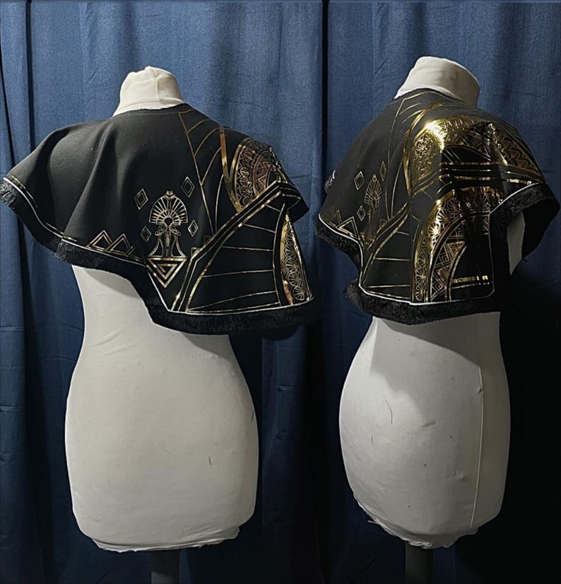 May include: Two black capes with gold geometric patterns and fringing are displayed on white mannequins. The capes have a short, rounded shape and are decorated with gold lines and shapes. The background is a dark blue curtain.