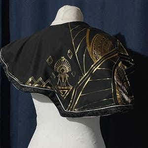 May include: Black cape with gold geometric patterns and trim. The cape has a white collar and is displayed on a mannequin. The gold designs include triangles, diamonds, and stylized sunburst patterns. The cape's edge is trimmed with a gold and black border.