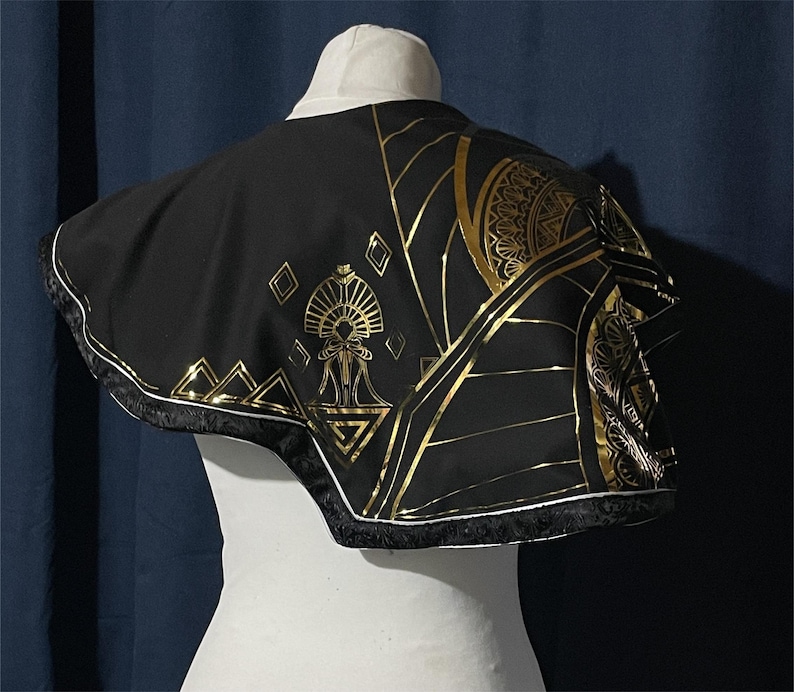May include: Black cape with gold geometric patterns and trim. The cape has a white collar and is displayed on a mannequin. The gold designs include triangles, diamonds, and stylized sunburst patterns. The cape's edge is trimmed with a gold and black border.
