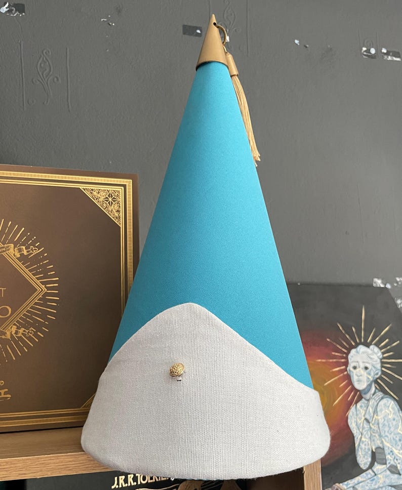 May include: A turquoise party hat with a white brim and a gold tassel. The hat has a gold tip and a small gold button on the front. The hat is made of felt and fabric.