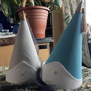 May include: Two cone-shaped hats, one gray and one teal, with white trim and gold accents. The gray hat has a sheer black veil. Both hats have a gold tassel and a gold button on the brim. The hats are on a floral patterned surface.