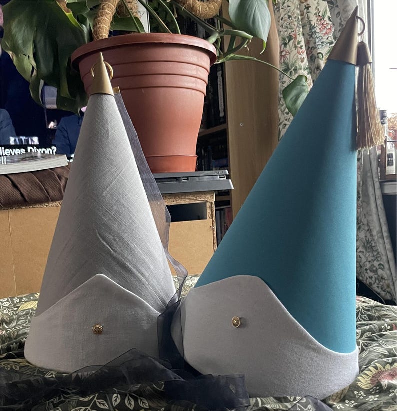 May include: Two cone-shaped hats, one gray and one teal, with white trim and gold accents. The gray hat has a sheer black veil. Both hats have a gold tassel and a gold button on the brim. The hats are on a floral patterned surface.
