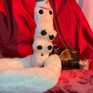 May include: White plush worm toy wearing a blue and green hat with a gold star. The worm is curled up on a red velvet surface.