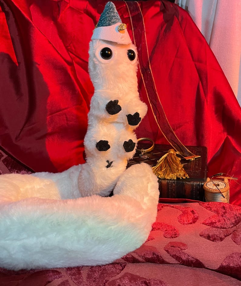 May include: White plush worm toy wearing a blue and green hat with a gold star. The worm is curled up on a red velvet surface.