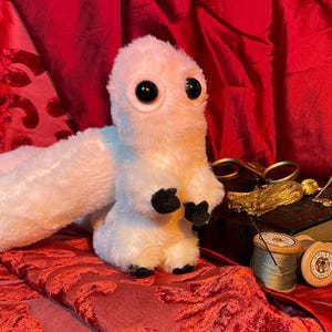May include: A white, fluffy, plush toy with large black eyes and a long tail. The toy is sitting on a red velvet surface.