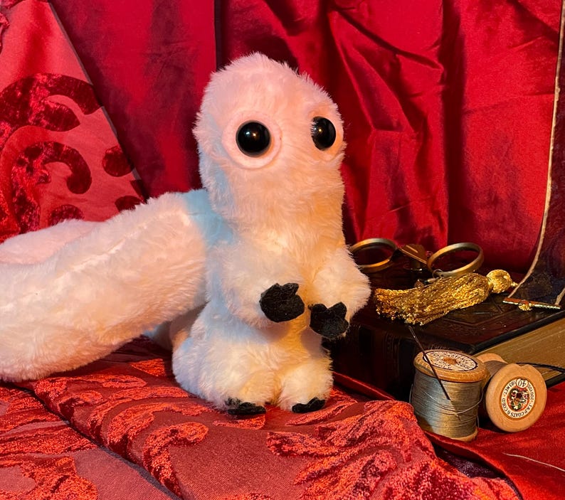 May include: A white, fluffy, plush toy with large black eyes and a long tail. The toy is sitting on a red velvet surface.