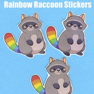 Könnte beinhalten: Drei Cartoon-Waschbär-Aufkleber mit grauen Körpern, dunkelgrauen Masken und regenbogenfarbenen Schwänzen. Die Aufkleber befinden sich auf einem hellblauen Hintergrund, mit dem Text "Rainbow Raccoon Stickers" oben.