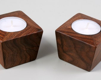 May include: Two wooden candle holders made of walnut wood. Each holder has a square base and a slightly angled top. The holders are empty and ready to hold tea lights.