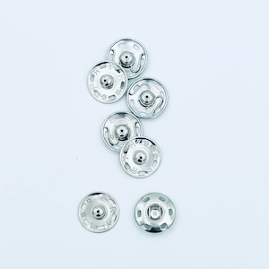 May include: A close-up of seven silver metal snap fasteners. The fasteners are round with a raised center and a decorative design around the edge.