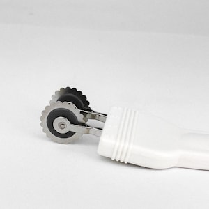 Op de afbeelding: Een witte plastic deegroller met twee zilveren wielen. De wielen hebben gekartelde randen en zijn ontworpen om deeg te snijden.