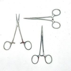 Pode incluir: Três pinças cirúrgicas prateadas com alças curvas e pontas pontiagudas. As pinças estão dispostas em forma triangular sobre um fundo branco.