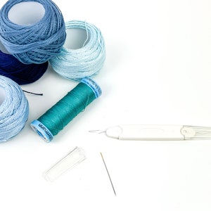 K&ouml;nnte beinhalten: Nahaufnahme einer Auswahl an blauen und gr&uuml;nen Stickgarnrollen, einem Nadelf&auml;der und einer Nadel. Die Garnrollen sind mit "DMC" und "Anchor" beschriftet.