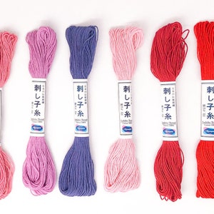 May include: A collection of six skeins of Olympus Sashiko Thread in various shades of pink, purple, and red. Each skein is labeled with Japanese text and product information. The cotton thread is tightly wound.