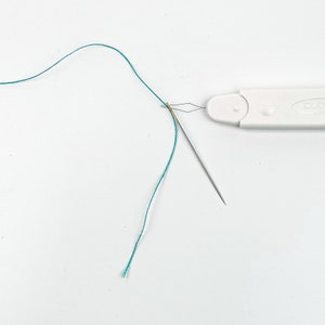 K&ouml;nnte beinhalten: Ein wei&szlig;er Plastik-Nadelf&auml;der mit einem gr&uuml;nen Faden, der durch die Nadel geht. Die Nadel ist gef&auml;delt und der Nadelf&auml;der liegt auf einer wei&szlig;en Oberfl&auml;che.