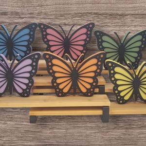 May include: A collection of six colorful butterfly decorations in shades of blue, pink, green, purple, orange, and yellow. Each butterfly has a black outline and is displayed on a small wooden shelf with black supports.