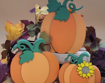 May include: Three wooden pumpkins in various sizes, painted in shades of orange. The largest pumpkin has a green stem and leaves. The smallest pumpkin has a green stem and leaves and a yellow flower.