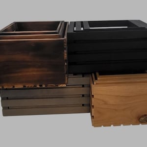 Könnte beinhalten: Eine Sammlung von Holzkisten in verschiedenen Farben, darunter Dunkelbraun, Schwarz, Hellbraun und Grau. Die Kisten sind gestapelt und zeigen ihre unterschiedlichen Größen und Oberflächen. Das Design weist eine Lamellenkonstruktion auf.