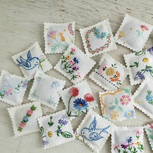 May include: A collection of 18 small, white, embroidered fabric squares with various floral and geometric designs. The squares are arranged in a random pattern on a white wooden surface.