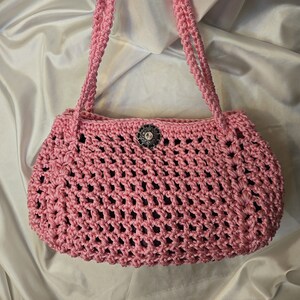 May include: A pink crocheted handbag with a decorative silver button. The bag has a woven design and two long straps.