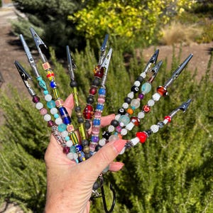 May include: A collection of beaded alligator clips in various colors and designs. The clips feature beads in shades of red, blue, green, and white, with some adorned with mushroom and other decorative elements. The clips are held together with a black clasp.