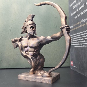 May include: A bronze-coloured sculpture of a muscular warrior wearing a crested helmet and holding a large bow. The figure is depicted from the waist up, posed in a dynamic stance with one arm extended to hold the bow, set on a rectangular base.