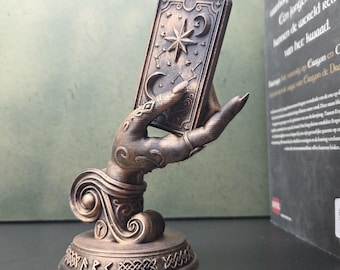 May include: A bronze-colored sculpture of a hand emerging from a swirling base, holding a rectangular card embossed with a star, crescent moon, and constellations. The circular base features engraved runic symbols and a decorative braided border.