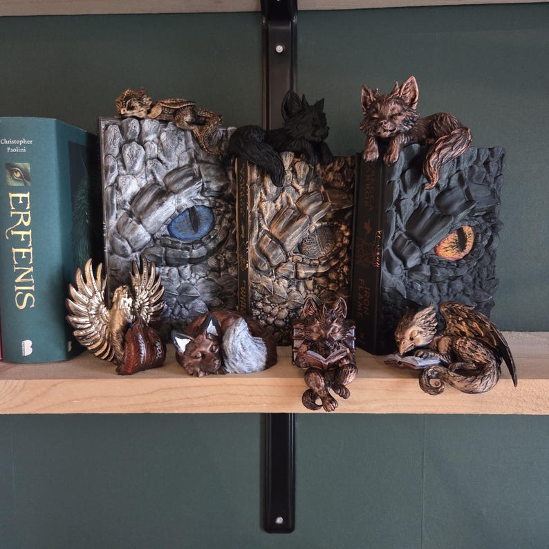 May include: A collection of fantasy-themed bookends and figurines. The bookends feature dragon and eye designs, while the figurines depict a fox, a griffin, and a reading fox. The color palette includes shades of brown, gray, and gold.