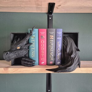 May include: Black dragon bookends with a detailed design, holding up a row of colorful books. The dragon's head and tail frame the books, creating a fantasy-themed display on a wooden shelf.