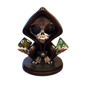 May include: A small, dark figurine of a Grim Reaper-like character with a skull face and hooded cloak. The figure holds two green and clear dice, one with a 0 and the other with a 3. The figurine is on a round base.