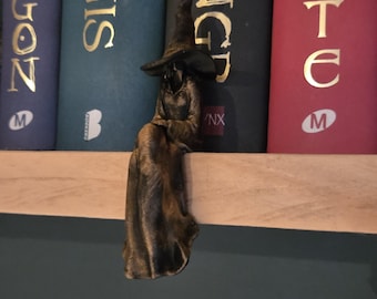 May include: A bronze-colored figurine of a seated witch wearing a pointed hat and a long, flowing robe. The figure is positioned with its legs dangling over the edge of a shelf, featuring detailed folds in the fabric and a dark, weathered finish.