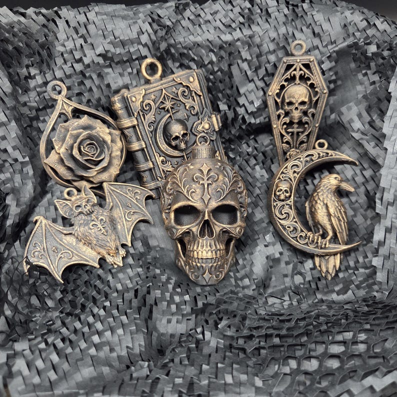 May include: A collection of five bronze-toned pendants featuring a rose, a book with a skull, a coffin with a skull, a bat, and a raven perched on a crescent moon. Each piece has a small loop at the top for hanging, showcasing intricate relief details.