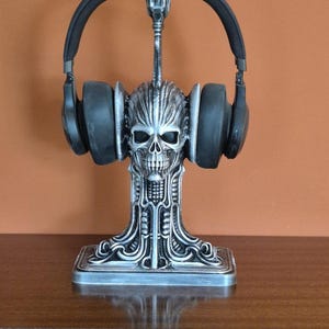 Könnte beinhalten: Silberfarbener Kopfhörerständer mit detailliertem Schädeldesign. Der Ständer hält ein Paar schwarze Kopfhörer. Der Sockel des Ständers ist quadratisch, das Design ist gotisch und künstlerisch.