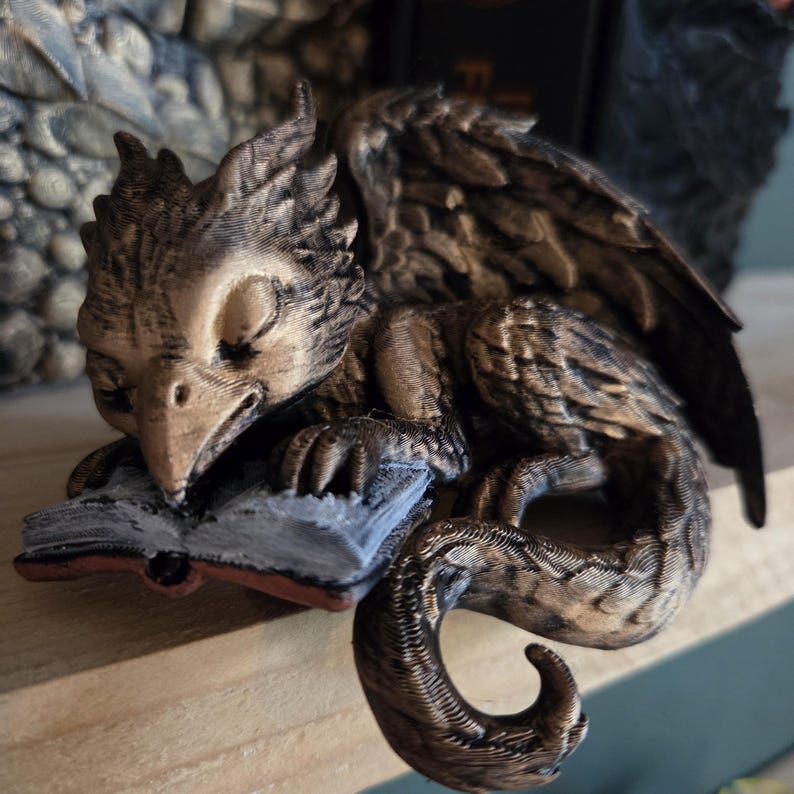 May include: A detailed figurine of a sleeping griffin, a mythical creature with the body of a lion and the head and wings of an eagle. The griffin is resting on a book with a red spine. The figurine is primarily brown and gold.