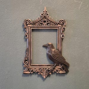 Puede incluir: Marco ornamental de color bronce con un diseño decorativo, con un cuervo posado. El marco tiene forma rectangular con intrincados adornos y una parte superior puntiaguda. El cuervo está detallado con plumas.