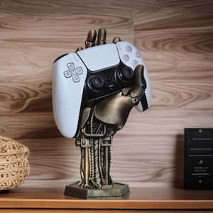 May include: A gold-coloured robotic hand holding a white PlayStation 5 controller. The hand is attached to a base with a mechanical design.