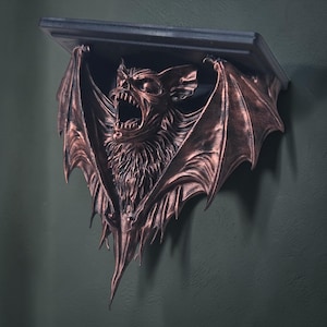 May include: A bronze-effect wall shelf with a detailed sculpture of a screaming bat. The bat has outstretched wings and a menacing expression. The shelf has a dark, rectangular top and is mounted on a dark green wall. The shelf is made of metal.