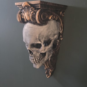 May include: A decorative wall shelf featuring a skull design. The skull is painted in shades of gray and white, with intricate details. The shelf has a gold and brown ornate bracket, adding a gothic touch. Perfect for displaying small items.