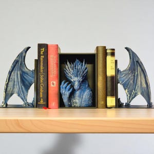 Könnte beinhalten: Drachen-Buchstützen-Set mit einer blauen Drachenskulptur in der Mitte. Die Buchstützen haben Drachenflügel auf jeder Seite. Bücher in verschiedenen Farben sind zwischen den Buchstützen platziert.